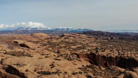 A-high-flying-drone-shot-over-the-vast-and-unique-desert-land-near-Moab,-Utah,-with-the-snowy-Rocky-Mountains-towering-in-the-distance