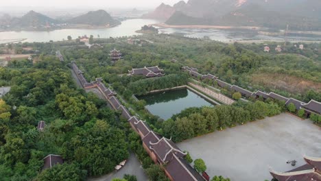 Drone-aerial-view-in-Vietnam-flying-over-a-buddhist-temple-area-filled-with-green-trees-in-front-of-a-serpent-river-in-Ninh-Binh-at-sunset