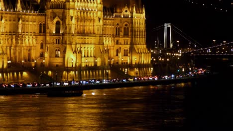 Vista-Del-Centro-De-La-Ciudad-De-Budapest-Con-El-Edificio-Del-Parlamento-Y-El-Río-Danubio-Por-La-Noche,-Arquitectura-Gótica,-Reflejos-De-Luz,-Toma-Panorámica-Distante