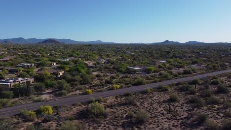 Cyclist-biking-on-long-desert-road-in-town-with-homes,-cactuses-and-mountains