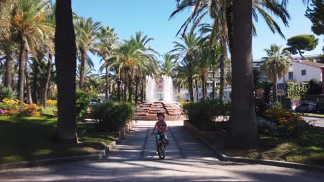 Young-boy-rides-bike-through-park-with-fountain-in-summer-in-Antibes