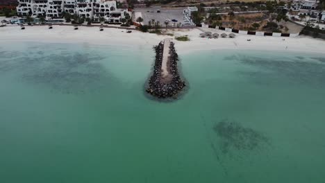 A-rocky-outcrop-in-turquoise-waters-on-beach,-playa-Caymancito,-near-La-Paz,-in-Baja-California-Sur-with-resorts-in-background,-aerial-view