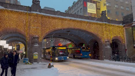 Bus-and-cars-pass-below-bright-cheerful-mat-of-lights-on-bridge-as-commuters-walk-on-snowy-covered-street-Kungsgatan-in-Stockholm,-Sweden