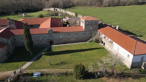 Tall-tree-casts-long-shadow-across-green-grass-and-walkway-of-monastery,-aerial-pan