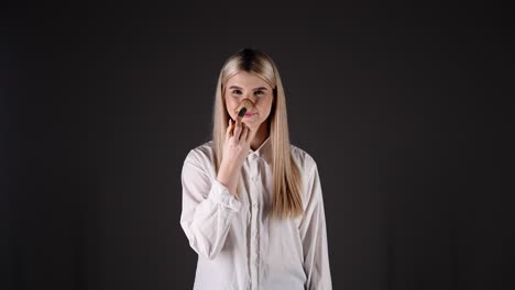 Mujer-Rubia-Sensual-Caucásica-Con-Camisa-Blanca-Juega-Con-Pincel-De-Maquillaje-Croma-Estudio-Negro-Fondo-Infinito,-Modelo-Joven-Rubia-Sonriendo