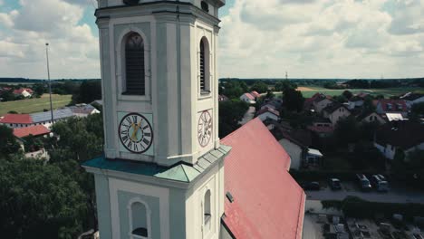 Ein-Uhrturm-Mit-Rotem-Dach-In-Einem-Malerischen-Dorf-An-Einem-Sonnigen-Tag,-Grüne-Landschaft-Im-Hintergrund,-Luftaufnahme