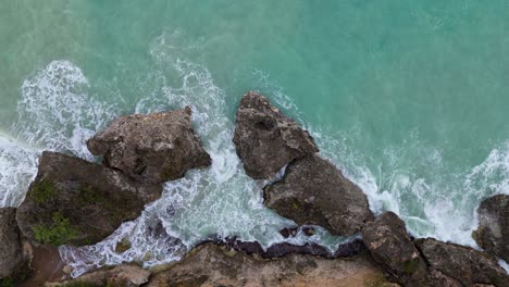 Aerial-descend-on-turbulent-water-spilling-between-coastal-rocky-boulders,-bird's-eye-view