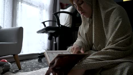 Young-Indian-female-Muslim-siting-cross-legged-quietly-reading-Holy-Quran-at-home
