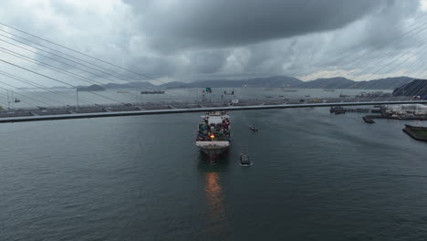 Container-vessel-approaching-under-stonecutter-suspension-bridge-cinematic-rising-drone-shot,-cloudy-sky