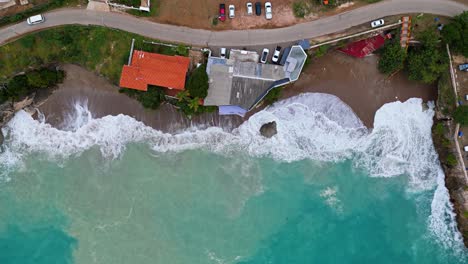 Bird's-eye-view-above-crashing-ocean-waves-churning-sand-along-coastal-home-on-highway-in-Caribbean-tropical-paradise