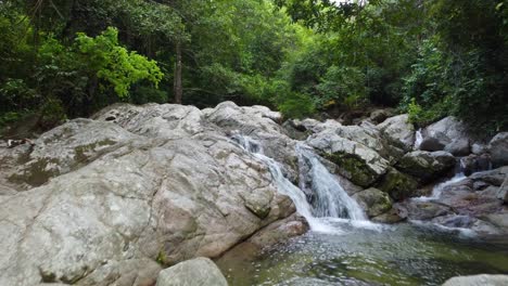 Waterfall-flowing-over-lush-dense-jungle-rainforest-rocky-stream-in-Minca-Columbia-moving-deeper-through-foliage