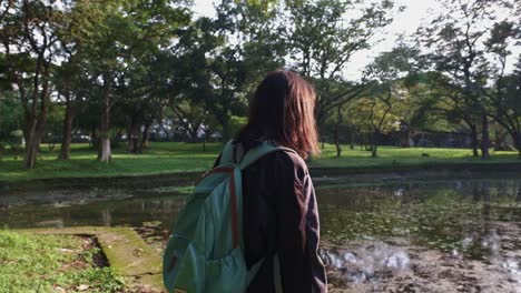 Young-Asian-Woman-With-Backpack-Walking-By-The-Pond-In-The-Park