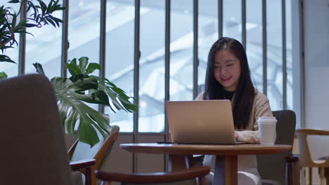 Joven-Asiática-De-Buen-Humor-Trabajando-Felizmente-En-Línea-En-Su-Computadora-Portátil-Desde-La-Cafetería