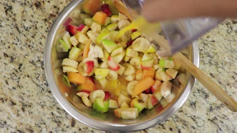 Top-shot-of-female-hand-pouring-fresh-orange-juice-into-freshly-chopped-fruit-bowl-on-a-kitchen-granite-slab