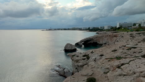 Brücke-Der-Liebe-Oder-Liebesbrücke-In-Zypern-In-Der-Dämmerung-Mit-Dramatischem-Himmel-Am-Kap-Greco,-Küste-Der-Stadt-Ayia-Napa-Im-Hintergrund---Luftaufnahme-Zurück-Zum-Rand-Der-Klippe