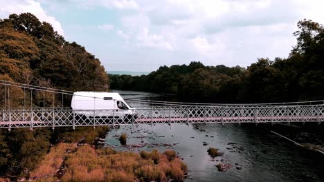 White-panel-van-driving-over-a-steel-suspension-bridge-spanning-a-river-in-Wales