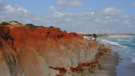 Aerial-view-of-the-sea,-waves,-cliffs,-a-small-village-and-a-wind-energy-at-background,-Morro-Branco,-Ceara,-Brazil