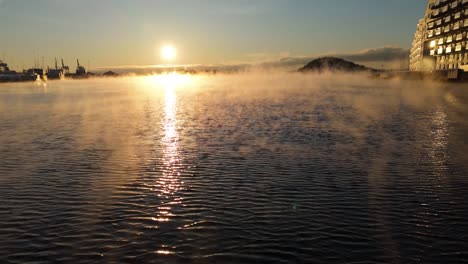 Schwebender-Nebel,-Der-Sich-über-Dem-Wasser-Neben-Wohnungen-Bewegt,-Mit-Goldenem-Sonnenuntergang-Im-Hintergrund-In-Bjorvika,-Oslo