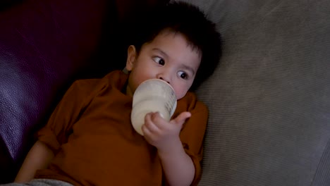 Adorable-18-month-old-baby-boy-sits-leisurely-on-a-home-sofa,-holding-a-bottle-of-milk-and-drinking-from-it,-bathed-in-the-warm-glow-of-TV-screen-light-reflections