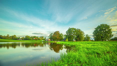 Golden-clouds-streak-across-blue-sky-reflecting-on-serene-pond-as-wind-blows-grass-and-trees-to-sway