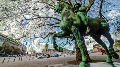 Toma-De-Timelapse-De-Una-Estatua-De-Un-Hombre-Montado-En-Una-Manguera-Con-Tráfico-Pasando-En-Hamburgo,-Alemania-Durante-El-Día