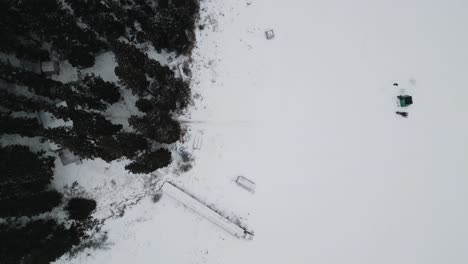 Eine-Drohnenaufnahme-Von-Oben-Einer-Insel-Auf-Dem-Zugefrorenen-Kanadischen-Paint-Lake-Mit-Einer-Eisfischerhütte-Und-Skioos