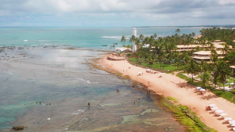 Aerial-view-of-Praia-do-Forte-beach,-the-coral-reef,-palm-tree-area-and-people-enjoying-the-sea,-Praia-do-Forte,-Bahia,-Brazil