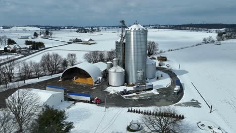 Toma-Aérea-Acercándose-A-Un-Silo-Industrial-En-Una-Granja-Con-Camiones-De-Transporte-En-Invierno