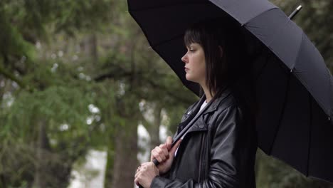 Portrait-of-the-worried,-nervous-young-woman-in-a-black-jacket-holding-the-black-umbrella-in-the-park