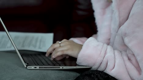 Hands-Of-Person-in-pink-robe-typing-on-laptop,-seated-indoors,-concept-of-work-from-home,-low-light,-cozy-atmosphere