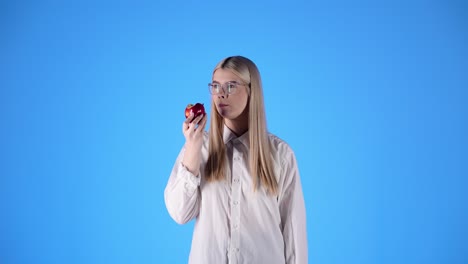 Cute-woman-takes-a-bite-from-tasty-red-organic-apple,-isolated-blue-background