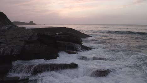 Drones-Volando-Sobre-Las-Olas-Del-Océano-Rompiendo-En-Las-Rocas-Al-Atardecer,-Aéreos-Hacia-Adelante-A-Baja-Altitud-Y-Copiando-Espacio