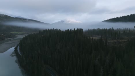 Misty-morning-over-Rovassaga-river-in-Norway,-with-forest-and-snow-capped-mountain