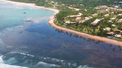 Aerial-view-of-Praia-do-Forte-beach,-the-coral-reef,-boats-parked,-palm-tree-area-on-a-cloudy-day,-Praia-do-Forte,-Bahia,-Brazil