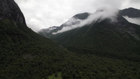Vista-Aérea-De-Montañas-Verdes-Y-Nubes-Que-Cubren-La-Cumbre-Del-Pico-En-Noruega-Con-Un-Estrecho-Curso-De-Agua-En-Cascada-Entre-Montañas