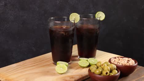Chilenischer-Piscola-Drink,-Typischer-Sommercocktail-Aus-Chile,-Glas-Mit-Eis,-Holzbrett,-Zitronen-Mit-Selektivem-Fokus,-Dunkler-Hintergrund