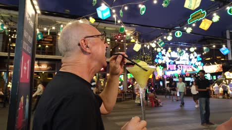 A-white-man-sipping-on-a-tall-glass-of-frozen-margarita-amidst-the-lively-and-crowded-evening-atmosphere-of-Pub-Street-in-Siem-Reap,-Cambodia