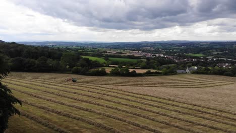 Aerial-panning-right-shot-of-a-tractor-gathering-hay-on-a-large-field-with-a-view-beyond-filmed-in-Devon-South-West-England