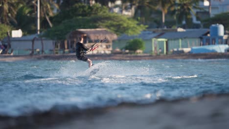Rear-view-of-kiteboarder-hopping-and-skipping-as-he-rises-to-do-360-spin,-blurred-background-of-home-and-coast-of-Dominican-Republic