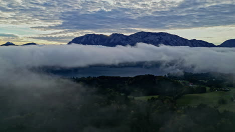 Panoramic-aerial-retreating-above-misty-thick-fog-or-cloud-layer-with-sunset-on-horizon-and-epic-snow-capped-mountain-ridgeline