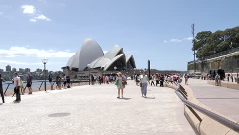 Wide-shot-of-people-walking-past-famous-Sydney-Opera-house-architectural-world-wonder-building-on-sunny-day,-New-South-Wales,-Australia