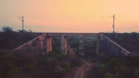 Aerial-drone-shot-of-an-Indian-railways-passenger-train-moving-fast-on-an-old-concrete-Railway-bridge-with-dense-forest-hills-in-background-during-late-evening-time