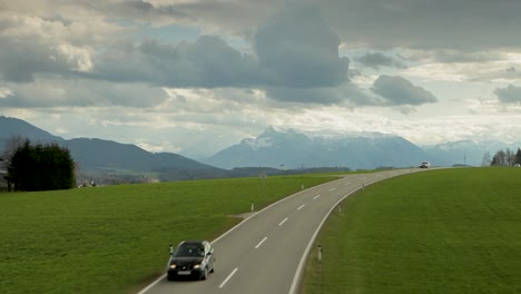 Los-Coches-Circulan-Por-Una-Carretera-Con-Exuberantes-Campos-Verdes-Y-Montañas-Cubiertas-De-Nieve-Como-Telón-De-Fondo,-Cielo-Parcialmente-Nublado-Arriba