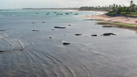 Vista-Aérea-De-Las-Rocas-En-El-Mar,-Los-Barcos-Estacionados,-área-De-Palmeras,-Olas-Y-La-Ciudad-Al-Fondo,-Guarajuba,-Bahía,-Brasil