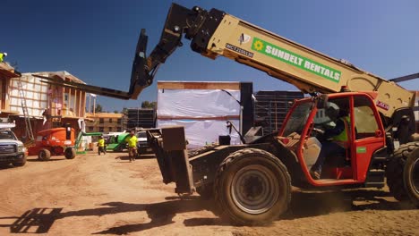 Gimbal-wide-panning-shot-of-a-construction-worker-operating-a-heavy-telehander-forklift-at-modular-building-site-in-West-Los-Angeles,-California