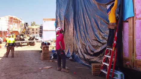 Gimbal-wide-shot-of-a-construction-crew-tarping-the-side-of-a-housing-module-at-a-pre-fab-building-site-in-West-Los-Angeles,-California