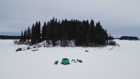 Una-Toma-De-Drone-De-Una-Isla-En-El-Lago-De-Pintura-Canadiense-Congelado-Con-Una-Cabaña-De-Pesca-En-Hielo-Y-Esquís