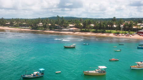 Aerial-view-of-Praia-do-Forte-beach,-the-coral-reef,-boats-parked,-palm-tree-area-on-a-cloudy-day,-Praia-do-Forte,-Bahia,-Brazil