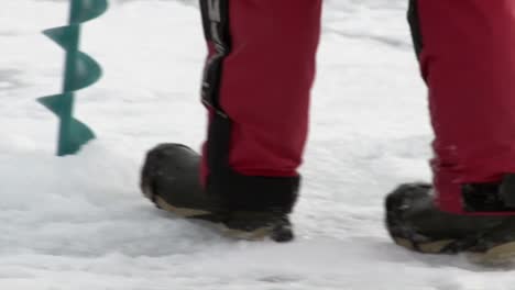 A-Man-in-Boots-Walking-on-Ice,-then-Using-an-Auger-to-Drill---Close-Up