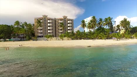 Bright-sunny-day-at-a-Miami-beach-with-clear-waters,-tall-palms-and-beachfront-buildings,-aerial-view,-in-America